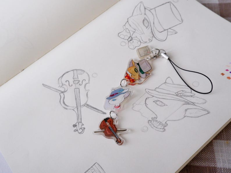 May include: A selection of cat-themed charms and a miniature violin pin are displayed on a sketchbook page with pencil sketches. The charms depict cats wearing hats and bow ties, with a clear, glossy finish. The violin pin is reddish-brown.