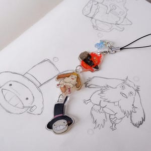 May include: A clear acrylic charm featuring four colourful cartoon characters from a fantasy film, connected by a silver chain and black loop. The charm is set against a white background with pencil sketches of similar characters.