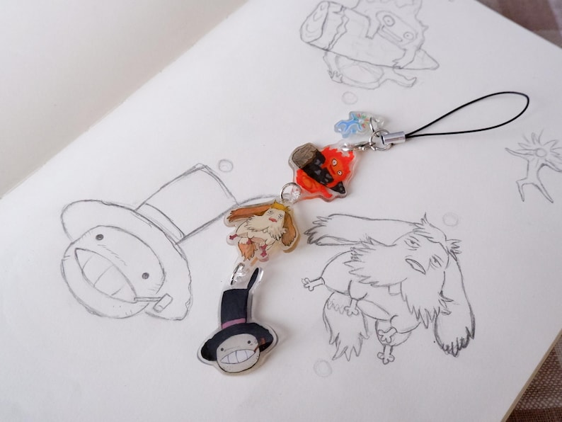 May include: A clear acrylic charm featuring four colourful cartoon characters from a fantasy film, connected by a silver chain and black loop. The charm is set against a white background with pencil sketches of similar characters.
