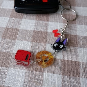 May include: Keychain featuring a black cat with purple ears, a red radio, a wreath, and two red bow charms. The charms are attached to a silver keyring. The keychain is a decorative accessory.