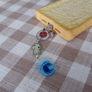 May include: A phone case designed to look like a slice of bread, attached to a charm with three anime-style characters. The charm includes a red and white mask, a small white creature, and a blue, stylized creature. The case rests on a brown and white checkered surface.