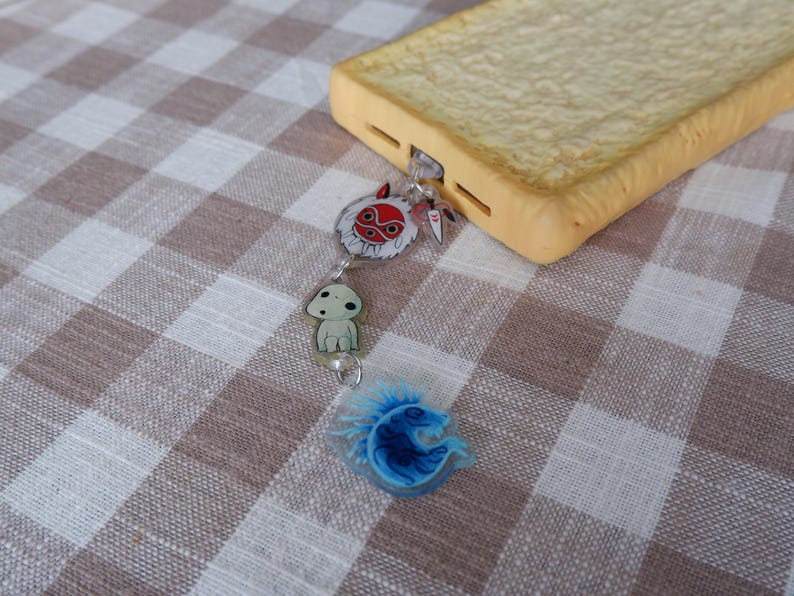 May include: A phone case designed to look like a slice of bread, attached to a charm with three anime-style characters. The charm includes a red and white mask, a small white creature, and a blue, stylized creature. The case rests on a brown and white checkered surface.