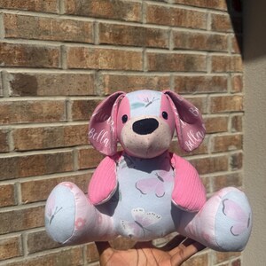 May include: A stuffed animal dog toy featuring a light blue fabric body with a pink and white butterfly pattern. The dog has pink ears with the name Bella printed in white script, a pink face, a black nose, and pink textured fabric arms.