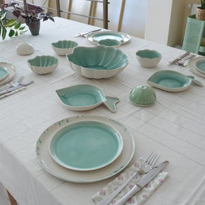 Op de afbeelding: Een tafelsetting met keramisch serviesgoed in wit- en tinten. De set bevat borden, kommen en serveerschalen in verschillende vormen, waaronder een visvormige schaal. Decoratieve zeepaardjes en bolvormige objecten zijn ook aanwezig.