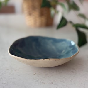 May include: A handmade ceramic bowl with a unique shape. The interior is a deep blue, contrasting with the off-white exterior. The bowl's rim has a slightly irregular edge, adding to its handcrafted appeal. The bowl is likely used for serving food or as a decorative piece.