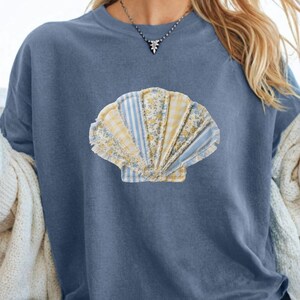 May include: A blue jean colored t-shirt featuring a seashell design. The seashell is a patchwork of various fabrics in shades of blue, yellow, and white. The shirt is a relaxed fit and has short sleeves. The text "Blue Jean" is visible in the upper right corner.