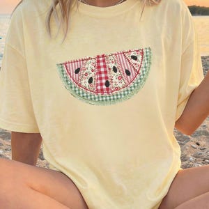 May include: Butter-colored t-shirt featuring a watermelon slice applique. The watermelon design includes red and white gingham, floral fabric, and black seed details. The shirt is a relaxed fit.