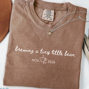May include: Espresso-colored t-shirt with the phrase "brewing a tiny little bean" and the date "NOV. 2026" printed in white. A silver necklace with a small pendant rests on the shirt. A brown hat is visible in the background.