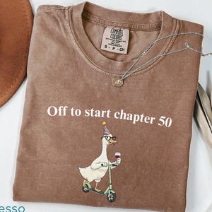 May include: Espresso-colored t-shirt with the text "Off to start chapter 50." A cartoon goose wearing glasses and a party hat is riding a scooter and holding a glass of wine. A silver necklace and a brown hat are also visible.
