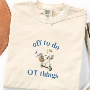 May include: An ivory-colored t-shirt featuring a graphic of a white goose riding a green scooter. The goose carries a brown bag filled with various tools and holds a coffee cup. The text above and below the graphic reads, off to do OT things.