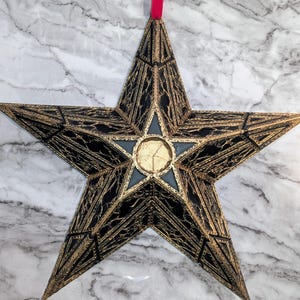 May include: A star-shaped ornament with a detailed geometric pattern. The gold-coloured frame has black and blue accents. A red ribbon is attached for hanging. The ornament is likely for festive or seasonal decoration, perhaps for Christmas.