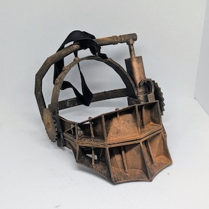 May include: A weathered, brown metal mask with a hinged jaw and a circular frame. The mask features sharp teeth and black straps for securing. This mask is a prop from a horror film.