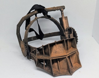 May include: A rusty brown metal mask with a hinged jaw and a circular frame. The mask has a series of sharp teeth and is secured with black straps. The mask is a prop from a horror movie.