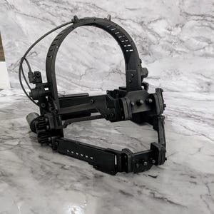 May include: A black metal head-worn device with a curved top and hinged jaw components. The device has multiple mechanical parts, including gears, bolts, and wires. It is set against a marble-patterned background.