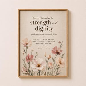 Könnte beinhalten: Gerahmter Druck mit hellbeigem Hintergrund. Der Text „Sie ist mit Stärke und Würde bekleidet“ ist zu sehen. Darunter blühen aquarellartige Blumen in Rosa und Weiß. Der Rahmen ist hellbraun. Der Druck enthält einen Bibelvers.
