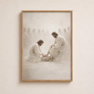 May include: A framed sepia-toned print depicts a biblical scene of two figures in white robes, one kneeling and washing the feet of the other. A group of figures are in the background. The artwork is encased in a light brown wooden frame.