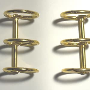 May include: Two gold-colored metal binder mechanisms, each featuring a central vertical spine attached to three circular rings. The rings are designed to open and close for holding paper, and the metal has a smooth, reflective, polished finish.