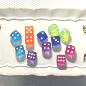 May include: A collection of colorful, translucent dice in various colors, including blue, orange, purple, green, and pink, are arranged on a white, decorative tray with gold bow handles. The dice are numbered with white dots.