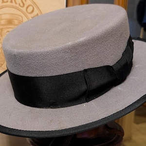 May include: A light grey fedora hat with a black grosgrain ribbon band and a black brim edge. The hat is displayed in front of a vintage sign that reads "Anderson Hats".