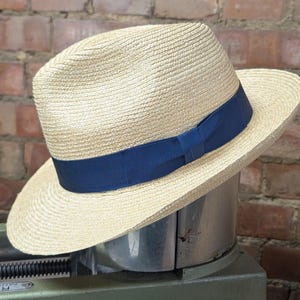 May include: A straw fedora hat with a light tan crown and brim. The hat features a dark blue grosgrain ribbon band. The hat is displayed on a metal surface against a brick wall background.