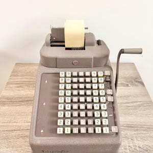 Op de afbeelding: Een vintage bruine mechanische rekenmachine met witte cijfertoetsen en een papieren rol. De rekenmachine heeft een handvat aan de zijkant en staat op een houten oppervlak. De toetsen zijn gerangschikt in rijen van cijfers van 1 tot 9.