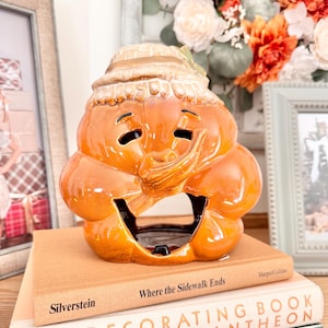 May include: A glossy orange ceramic pumpkin figurine featuring a carved face with a wide, open mouth and a textured hat on top. The pumpkin has a hollow interior and is designed as a decorative piece with a smooth, reflective finish and dark accents.
