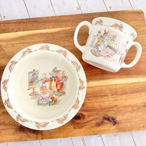 Puede incluir: Un juego de cuenco y taza de cerámica color crema con temática de conejos. El cuenco presenta un borde de conejos corriendo y una ilustración central de conejos pintando. La taza tiene dos asas y una escena de conejos similar. El juego está sobre una superficie de madera.