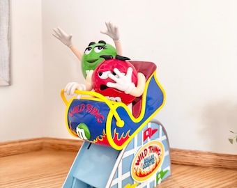May include: A plastic M&M's Wild Thing roller coaster toy featuring a red character and a green character riding in a blue and yellow car. The base is light blue with a white grid pattern and a circular logo that reads Wild Thing Roller Coaster.