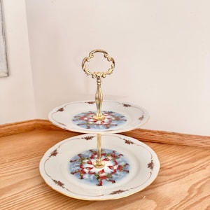 May include: A two-tier serving stand featuring white porcelain plates with gold-toned rims and a central gold-colored metal handle. The plates are decorated with a circular pattern of small figures wearing red hats and green clothing on a blue background.