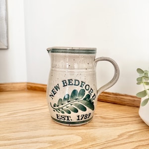 Puede incluir: Jarra de cerámica moteada con asa. La jarra tiene una base beige claro con un borde y asa verde oscuro. Las palabras "NEW BEDFORD" y "EST. 1787" están impresas en verde oscuro con un diseño de hojas.