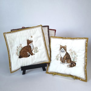May include: Two square quilted fabric panels featuring embroidered brown and white cats sitting among small floral stems. The panels have white centers, mustard yellow borders with white floral patterns, and decorative stitching along the edges.