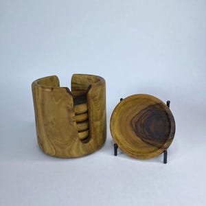 May include: A set of wooden coasters and a holder. The coasters are round with a dark brown center and light brown outer ring. The holder is a cylindrical shape with a cutout for the coasters. The wood grain is visible.