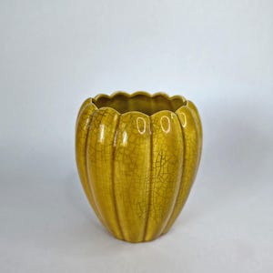 May include: A mustard yellow ceramic vase with a scalloped, pumpkin-like shape and a distinct crackle glaze finish. The vessel features vertical ridges running from the base to the wavy rim, creating a textured appearance across the surface.