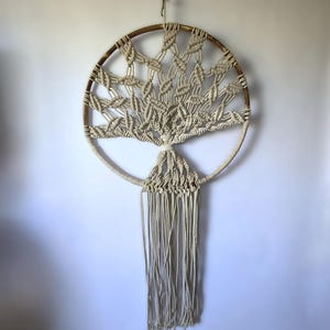 May include: A beige macrame wall hanging featuring a circular design with a tree-like pattern. The macrame is made of woven cotton rope and hangs from a wooden hoop. The design includes a fringe of hanging cords.
