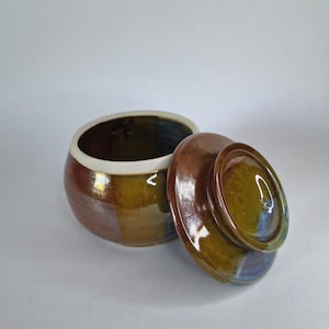 May include: A handmade ceramic jar with a lid, featuring a blend of brown, olive green, and blue hues. The jar has a white rim and a glossy finish. The lid is partially open, revealing the interior.