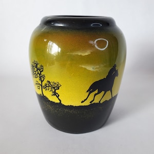 May include: A decorative ceramic vase with a black rim and base, featuring a yellow and orange gradient. The vase is adorned with a black silhouette of a horse and rider, trees, and a moon, creating a landscape scene.