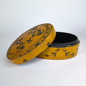 May include: Oval-shaped yellow box with a black interior and a lid. The lid is open, revealing the inside of the box. The box and lid are decorated with black floral patterns. The box is likely made of a leather-like material.