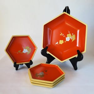 May include: Hexagonal red serving trays with gold trim and floral designs. The trays are displayed on black stands. The floral design includes white and gold blossoms. A set of four trays are stacked.