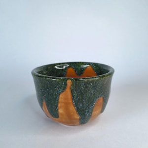May include: A small ceramic bowl featuring a smooth, warm terracotta-colored base contrasted by a thick, dark forest green glaze that drips down from the rim in a series of vertical, textured streaks, creating a layered and organic appearance.