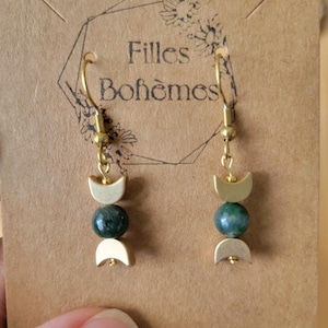 Peut inclure: Boucles d'oreilles dorées avec un motif de croissant de lune et une perle vert foncé. Les boucles d'oreilles sont suspendues à une carte marron avec le texte "Filles Bohèmes". Fermeture à crochet.