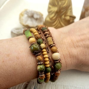 May include: A multi-strand bracelet featuring a combination of green, tan, and brown beads made of stone and wood, interspersed with copper-toned metal spacers and a copper-toned chain link element, creating a layered and textured appearance.