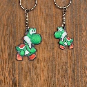 May include: Two keychains featuring a pixelated green dinosaur character with red and white accents. Each keychain has a silver chain and ring attached to a circular metal piece. The keychains are displayed against a brown wooden background.