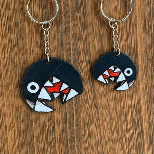 May include: Two black keychains with a cartoonish design of a mouth with white teeth and red accents. Each keychain has a silver-colored ring and chain. The keychains are hanging against a brown wooden background.
