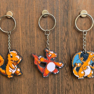 Puede incluir: Tres llaveros de personajes pixelados con cadenas y anillos plateados. Los personajes son naranjas, rojos y azules, con detalles blancos. Los llaveros cuelgan de una superficie de madera.