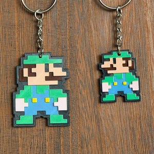 May include: Two keyrings featuring pixelated green and blue characters, each with a silver keyring and chain. The characters are wearing blue dungarees, green shirts, and brown hats. The keyrings are on a wooden surface.