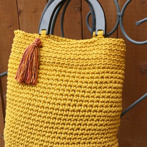 May include: A mustard yellow crocheted tote bag featuring a textured stitch pattern, two semi-circular black wooden handles, and a decorative multi-colored tassel attached to one side of the handle base, creating a functional accessory.