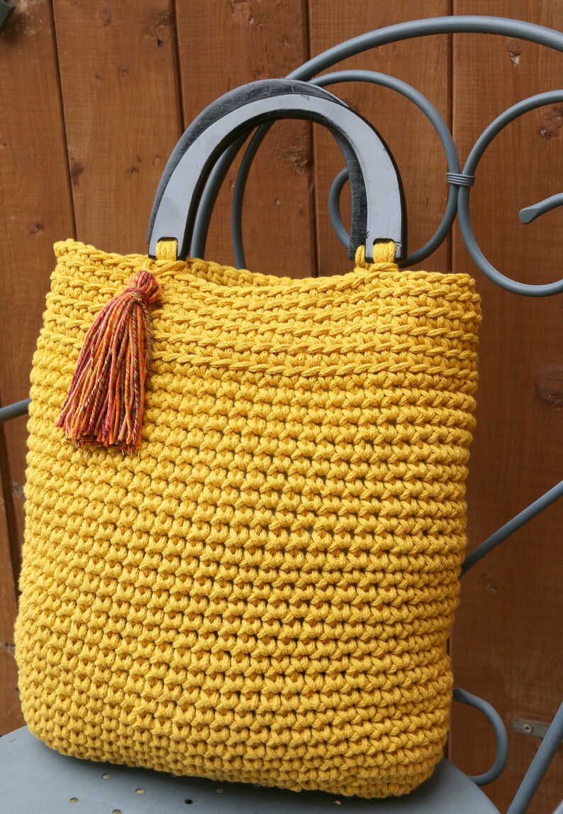 May include: A mustard yellow crocheted tote bag featuring a textured stitch pattern, two semi-circular black wooden handles, and a decorative multi-colored tassel attached to one side of the handle base, creating a functional accessory.