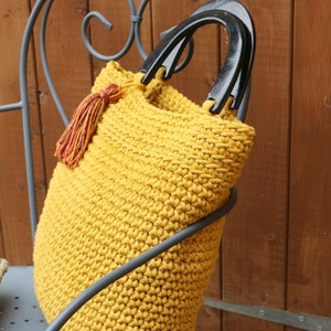 May include: A mustard yellow crocheted handbag featuring a textured stitch pattern, two glossy black semi-circular handles, and a decorative multi-colored tassel in shades of orange, red, and yellow attached to the side of the bag opening.