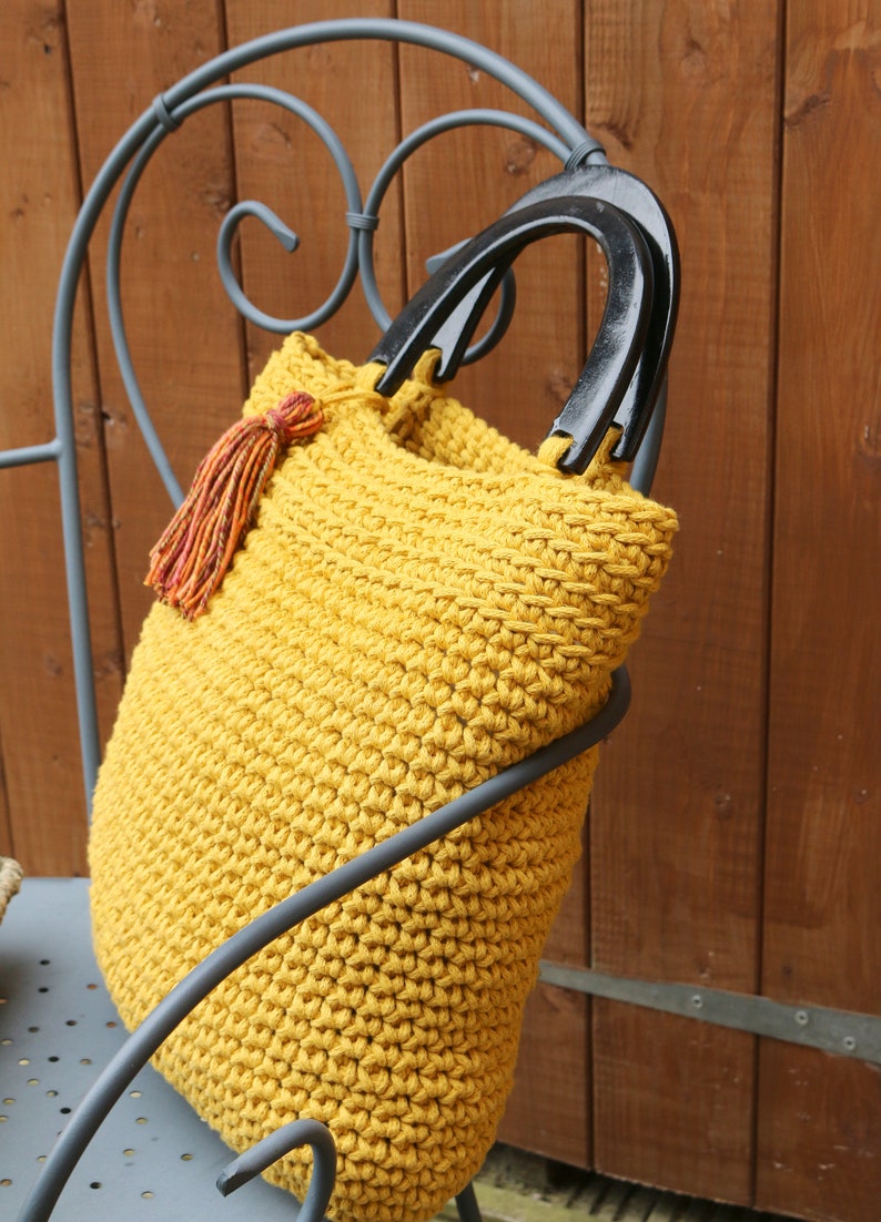 May include: A mustard yellow crocheted handbag featuring a textured stitch pattern, two glossy black semi-circular handles, and a decorative multi-colored tassel in shades of orange, red, and yellow attached to the side of the bag opening.