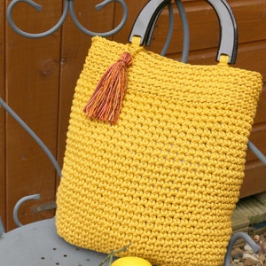 May include: A mustard yellow crocheted handbag featuring a textured stitch pattern, a dark brown semi-circular handle, and a decorative multi-colored tassel attached to the top. The bag has a square shape and is crafted from thick yarn.